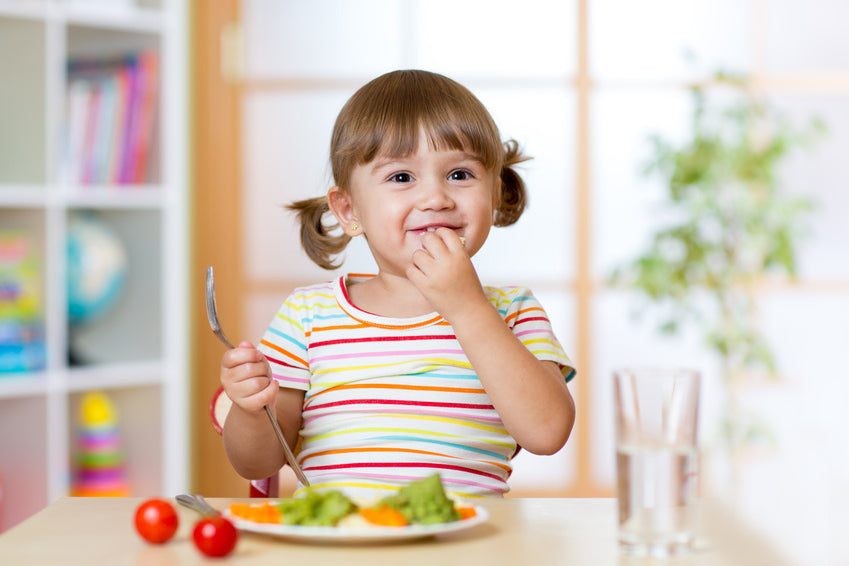 Toddler girl sitting at table eating vegetables and smiling