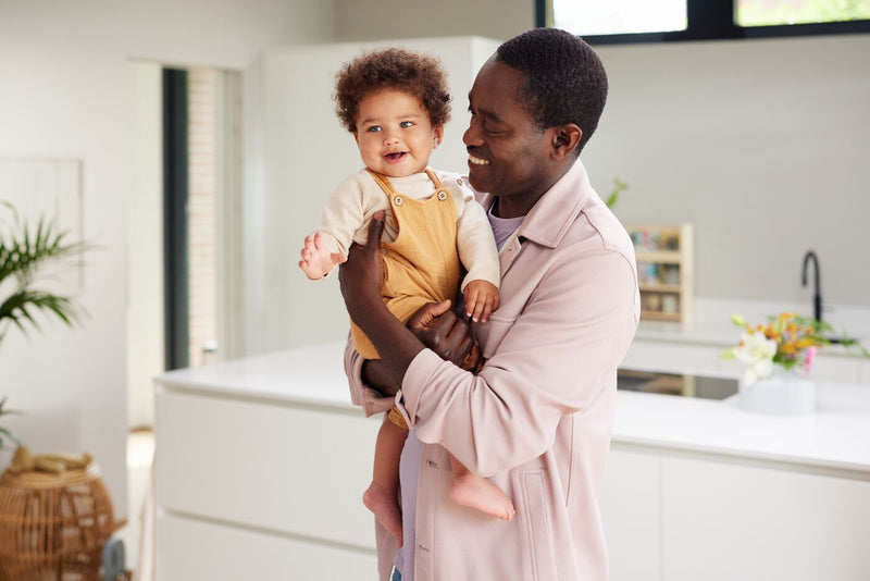 Dad in a pink shirt smiling and holding child in yellow overalls