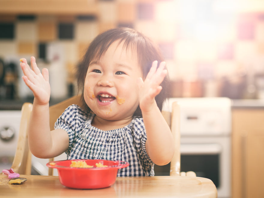 Smiling toddler eating meal from bowl with hands raised in excitement