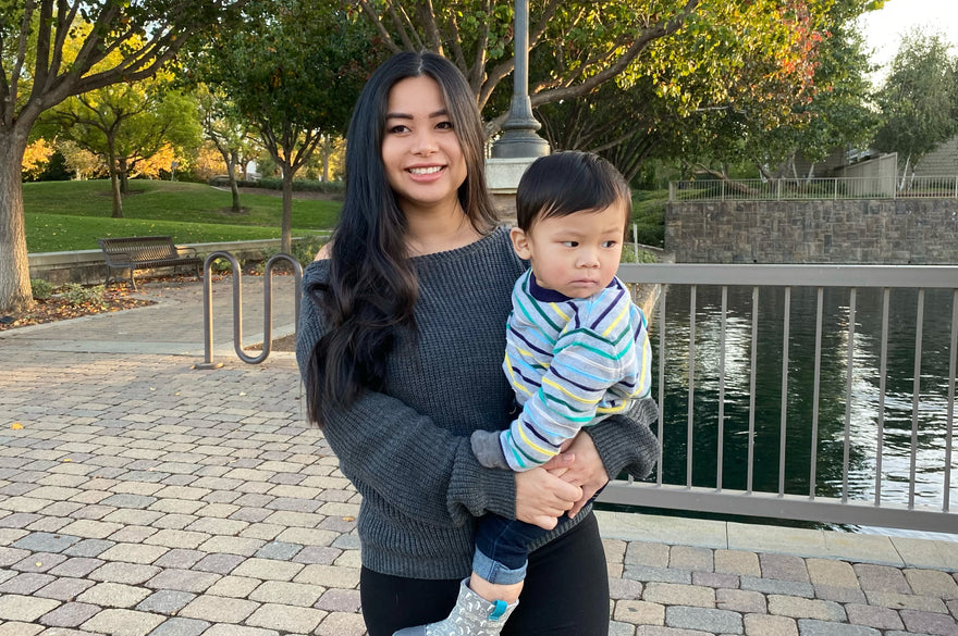Pricilla smiling holding her son Leo in a sunny park with water behind them.